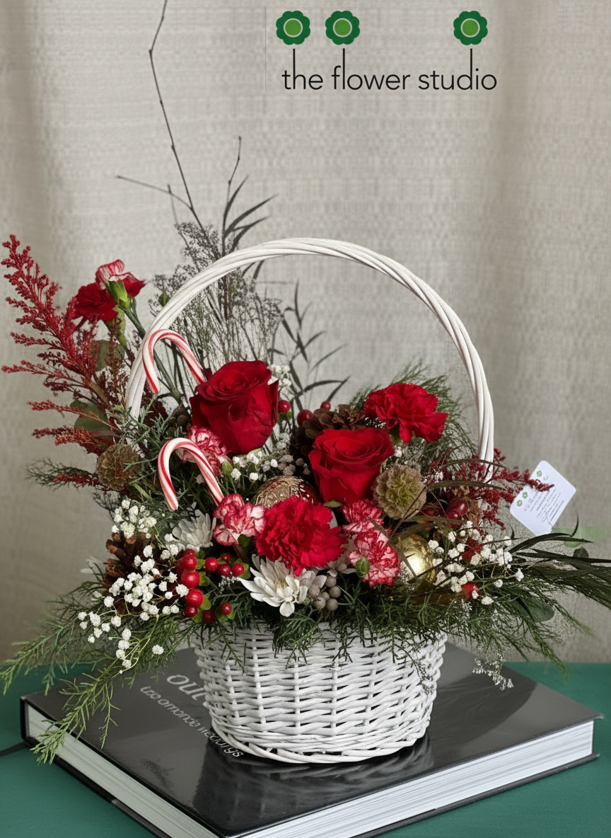 A decorative basket featuring red roses, greenery, white accents, and candy canes, displayed on a table with a studio backdrop.