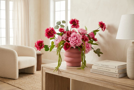 A pink ceramic flower vase filled with a bouquet of artificial pink roses, peonies, and green eucalyptus stems, displayed on a green tufted velvet surface against a beige curtain backdrop.