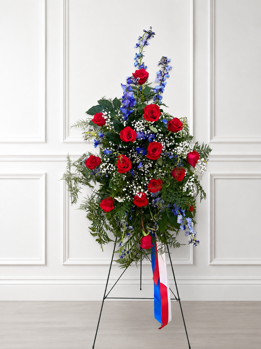 A large floral arrangement featuring red roses, blue delphiniums, and green foliage on a black metal stand, with a red, white, and blue ribbon hanging from the base, set against a white paneled wall.