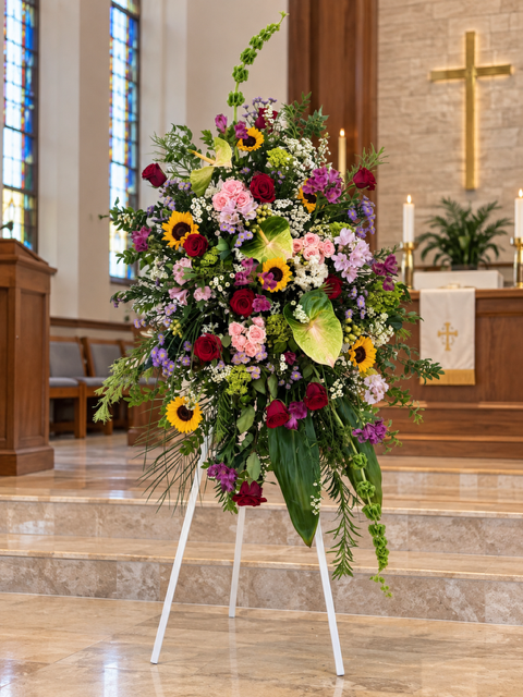 Colorful Farewell standing sympathy spray featuring sunflowers, deep red roses, soft pink flowers, purple blooms, and cascading greenery. Displayed on a white easel inside a church. Handcrafted by The Flower Studio Oxford, serving The Villages, Florida.