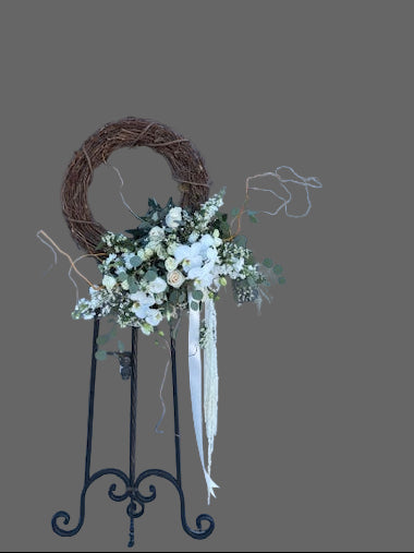 Decorative wreath with white flowers and greenery on a stand against a gray wall.