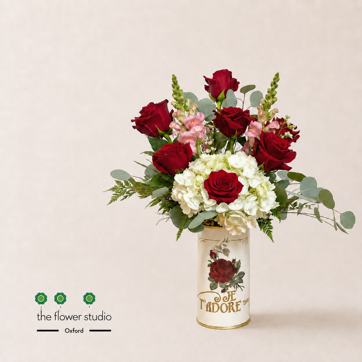 A floral arrangement featuring deep red roses, white hydrangeas, and pink carnations with green foliage, presented in a decorative cream-colored cylindrical vase printed with 'Je T'Adore' and a rose illustration. The arrangement is photographed on a light beige background with 'the flower studio' and 'Oxford' branding in the lower-left corner.