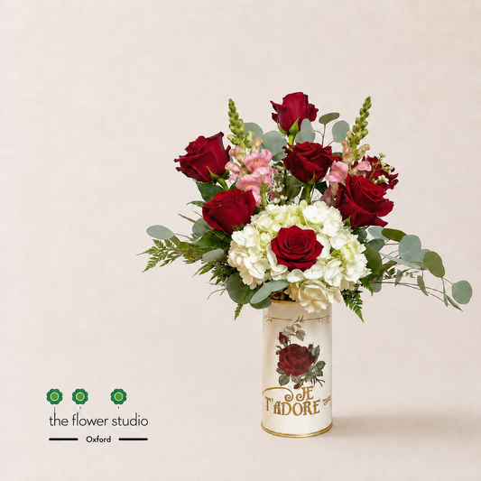 A floral arrangement featuring deep red roses, white hydrangeas, and pink carnations with green foliage, presented in a decorative cream-colored cylindrical vase printed with 'Je T'Adore' and a rose illustration. The arrangement is photographed on a light beige background with 'the flower studio' and 'Oxford' branding in the lower-left corner.